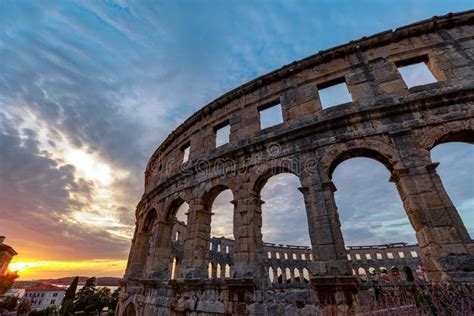 Mesa imperial romana frente al Coliseo recién construido, enmarcado por casas de piedra y vegetación mediterránea al atardecer.